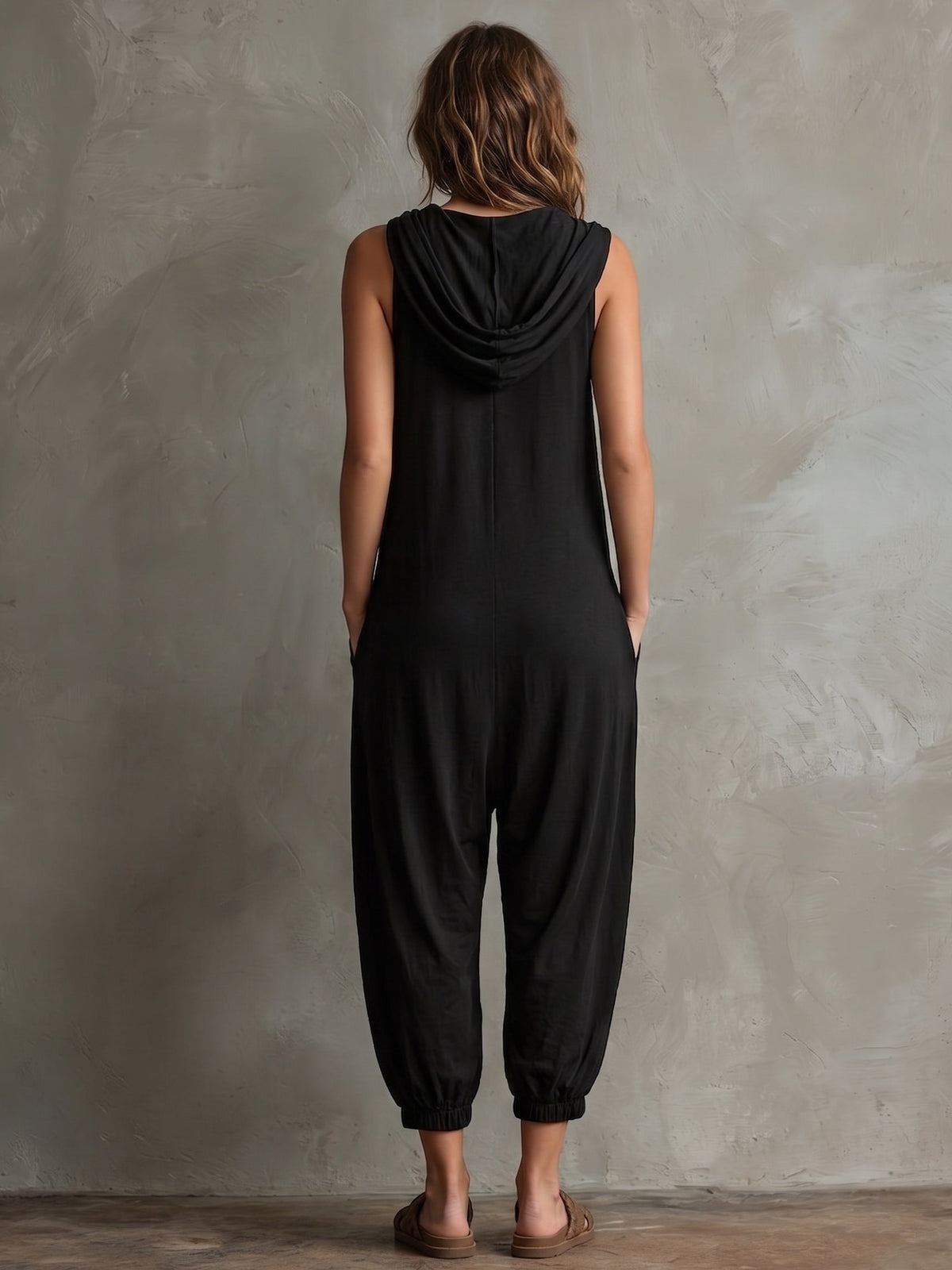 Black hooded sleeveless jumpsuit, rear view, with loose lantern legs and gathered waist